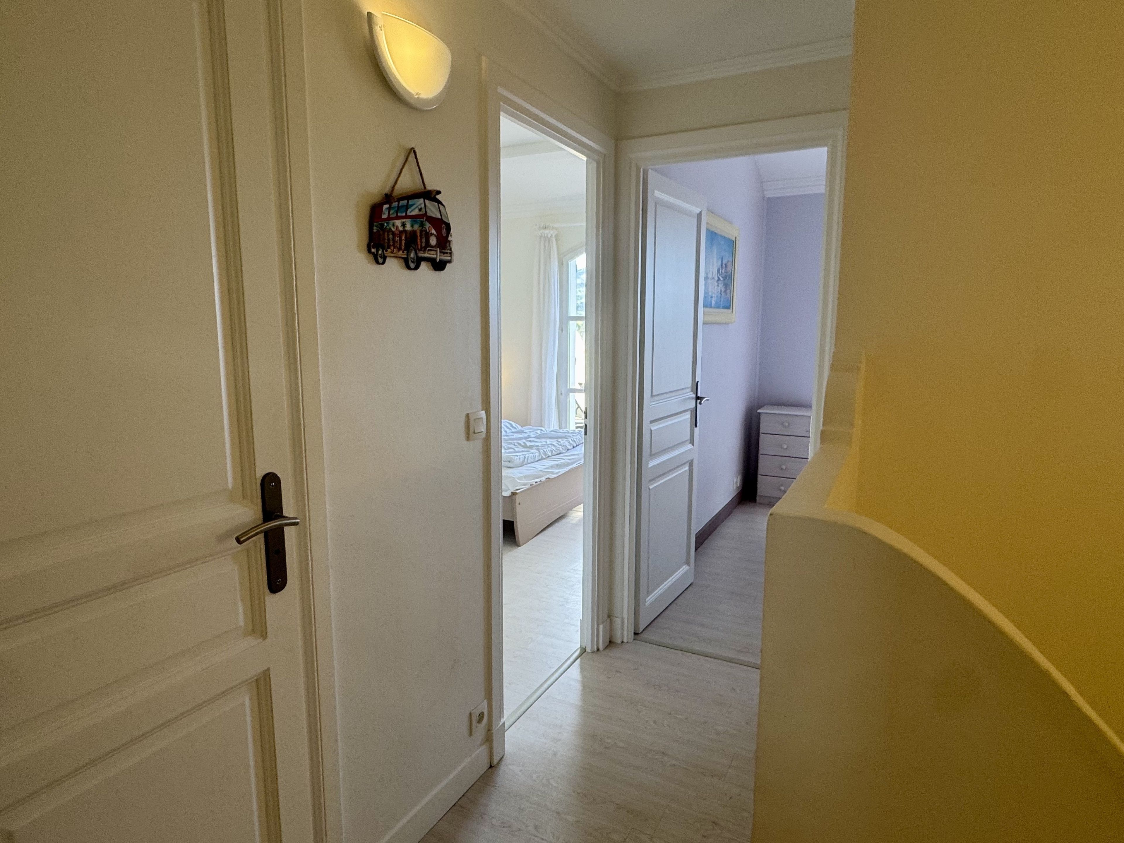 A hallway with a bed in the background, a dresser, and a wall-mounted light fixture.