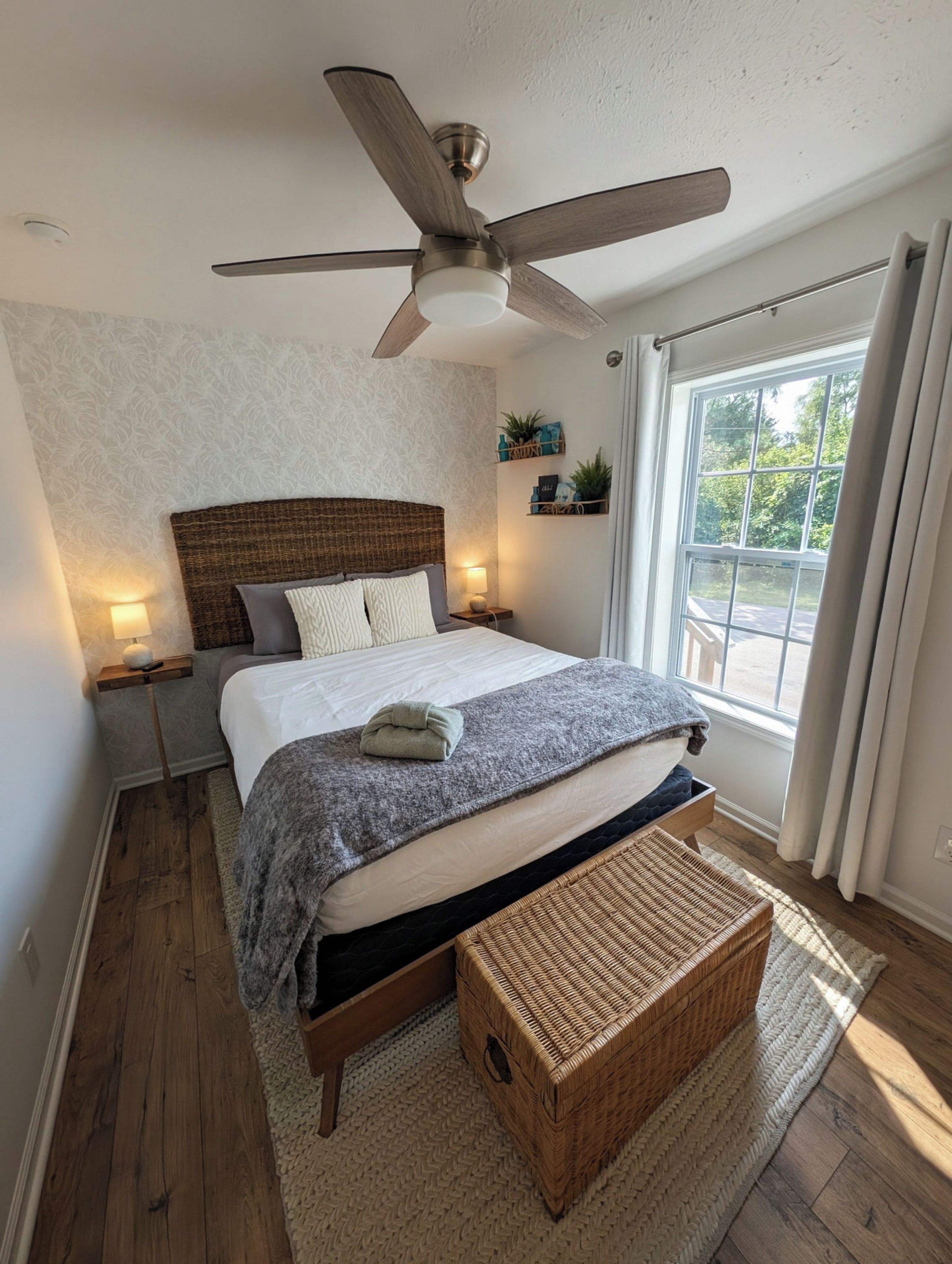 Een slaapkamer met een bed, een plafondventilator, een raam met gordijnen, een nachtkastje en een bijzettafeltje.