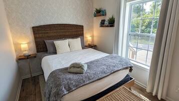 Een slaapkamer met een bed, een plafondventilator, een raam met gordijnen, een nachtkastje en een bijzettafeltje.