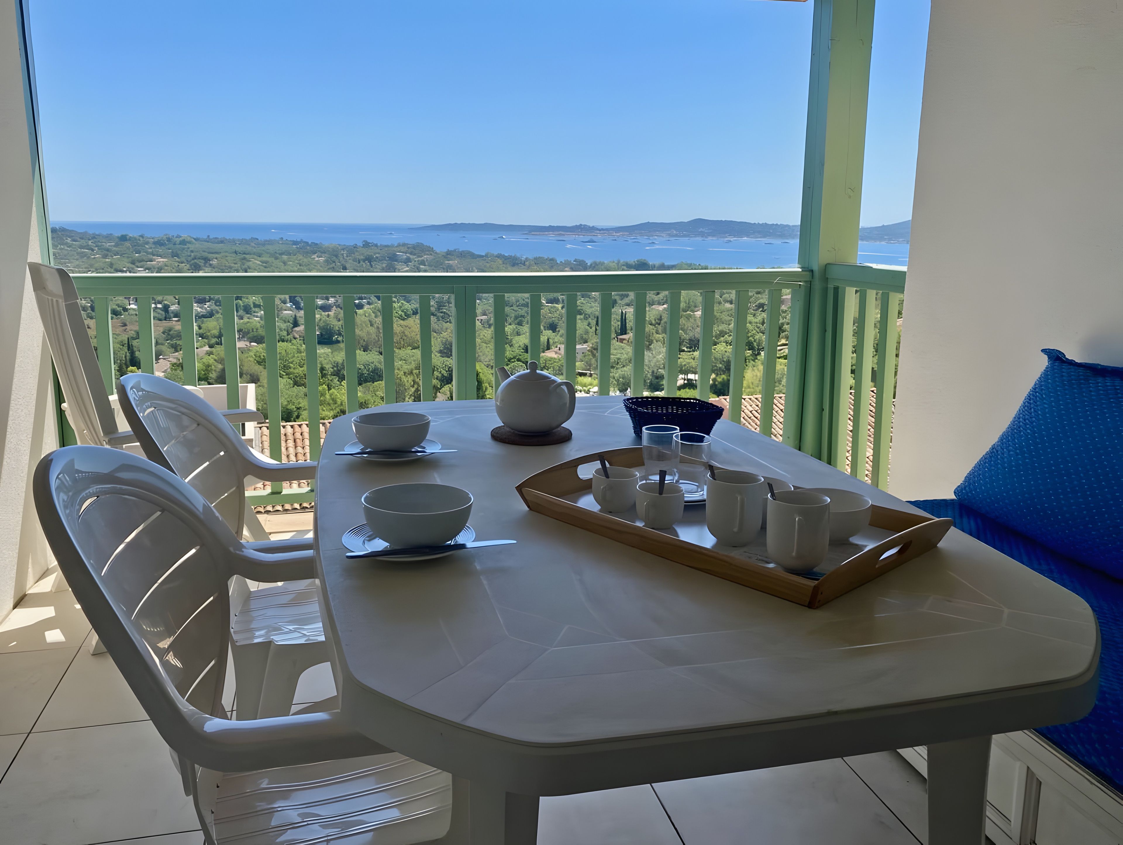 A balcony with a table set for dining, chairs, a view of the sea, and a green railing.