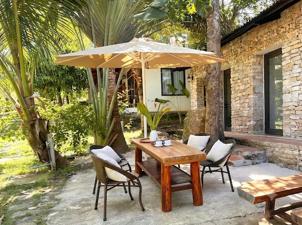 An outdoor seating area with a wooden table and chairs, an umbrella, and a stone building in the background. - Cay Sao Hill Resort Phu Quoc (Phu Quoc)