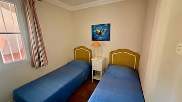 Un dormitorio con dos camas individuales, una mesita de noche con lámpara, un cuadro en la pared y una ventana con cortinas.