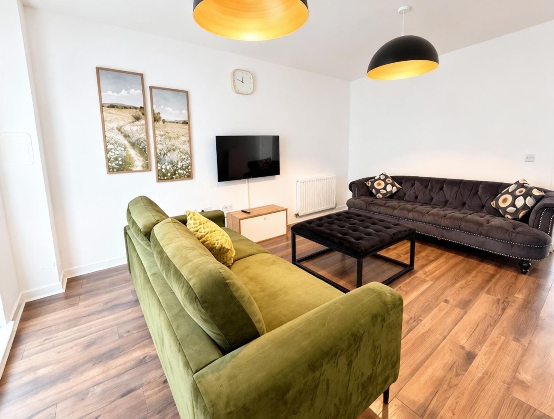 A modern living room with a green sofa, a black leather couch, and a coffee table. There is a flat-screen TV mounted on the wall and a clock.