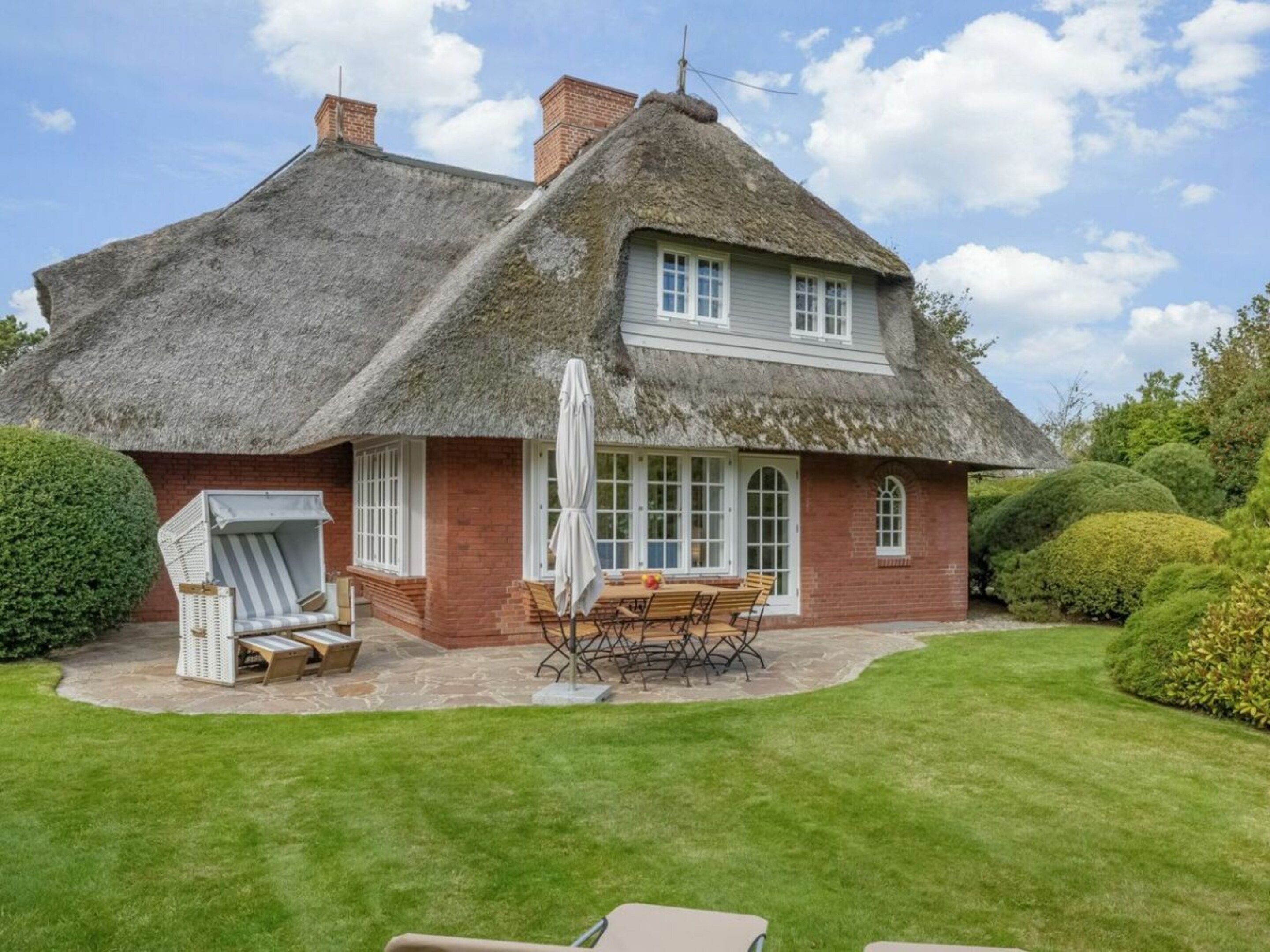 Une maison à toit de chaume avec une façade en briques, une terrasse avec une table et des chaises, et une pelouse soigneusement entretenue.
