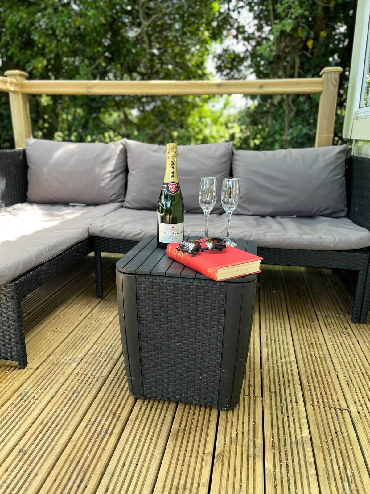Een rieten bank met grijze kussens, een zwarte tafel met een fles champagne, twee glazen en een rood boek.