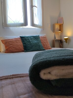 A bed with white bedding and multiple pillows, a bedside table with a lamp, and a window with sheer curtains.