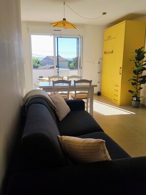 A living room with a sofa, a dining table with chairs, a yellow wardrobe, and a potted plant.