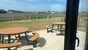An outdoor patio area with round wooden tables and benches, a view of a field, and a distant building. - 2-bedroom rental stay in peaceful Langemark-Poelkapelle (Langemark-Poelkapelle)