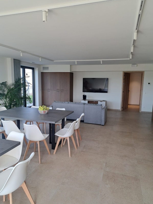 Un espacio habitable moderno con comedor, un sofá gris, televisor de pantalla plana y un ventanal grande.