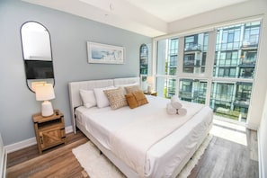 Un dormitorio con una cama grande, una mesita de noche, una lámpara, un cuadro enmarcado y una ventana con vistas a edificios.