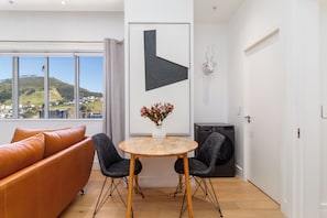 A modern living room with a sofa, a round table, and chairs, a large window with a view of a hill, and a door with a wall-mounted light fixture. - Luxury City Escape | Views and City Centre (Cape Town)