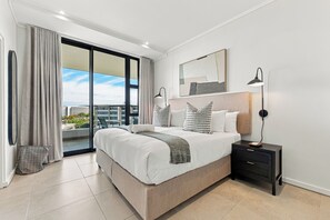 Quarto moderno com uma cama grande, mesa de cabeceira, luminária e vista da cidade através de uma varanda.