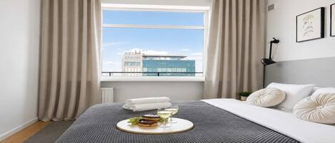 A hotel room with a large bed, a window with curtains, a view of a building, and a tray with food and drinks.