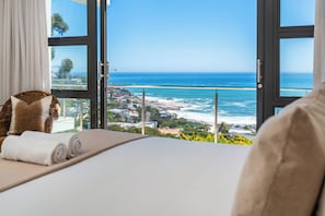 Ein Schlafzimmer mit großem Fenster und Blick auf die Küste, ein Bett mit gerollten Handtüchern und ein Korbstuhl.