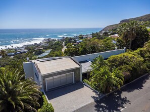 A coastal residential area with a modern house featuring a garage, surrounded by lush greenery and a view of the ocean. - Luxury Llundudno Villa | Private Pool and Garden (Cape Town)
