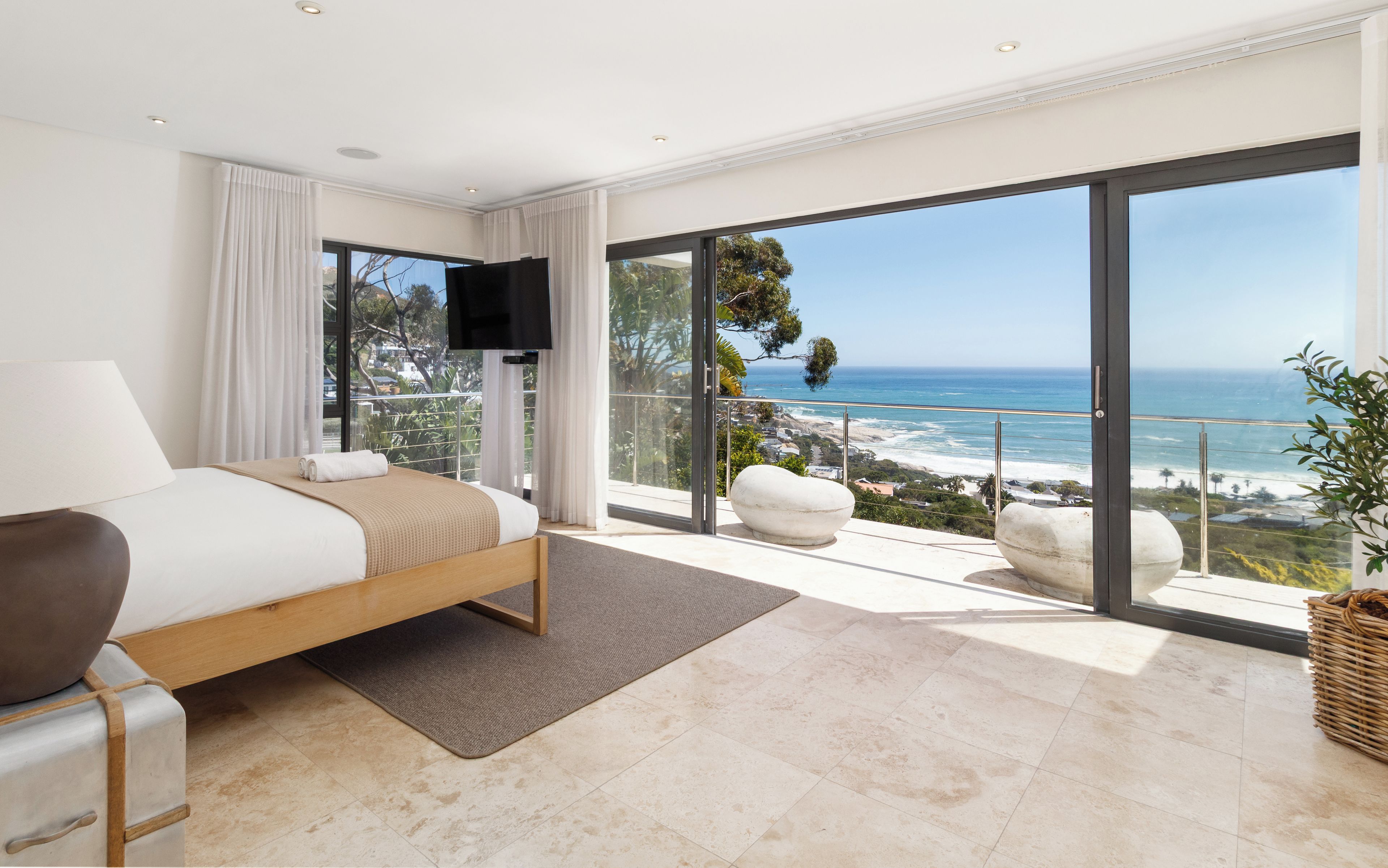 A bedroom with a bed, a flat-screen TV, a large window with a view of the ocean, and a balcony with a white chair.