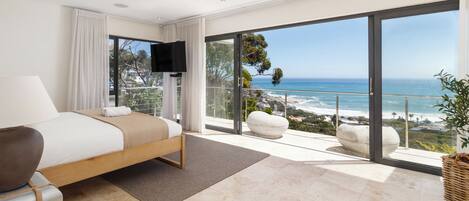 A bedroom with a bed, a flat-screen TV, a large window with a view of the ocean, and a balcony with a white chair.
