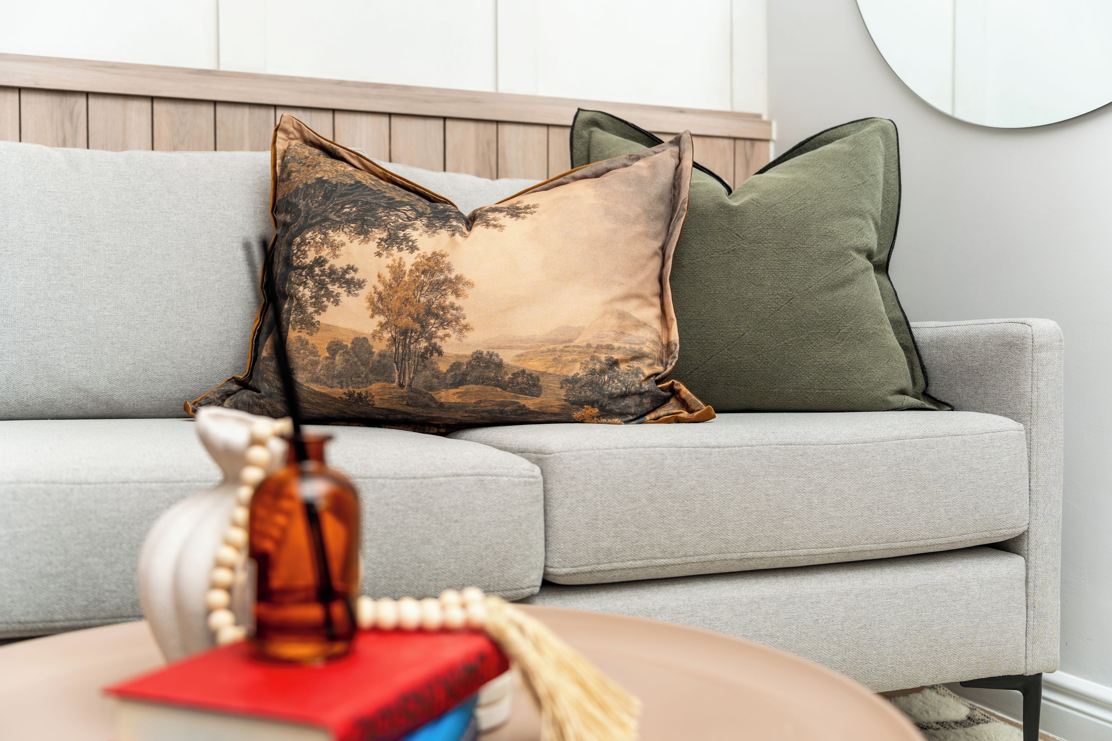 A grey sofa with two decorative pillows, a small table with a book and a vase, and a wooden headboard.