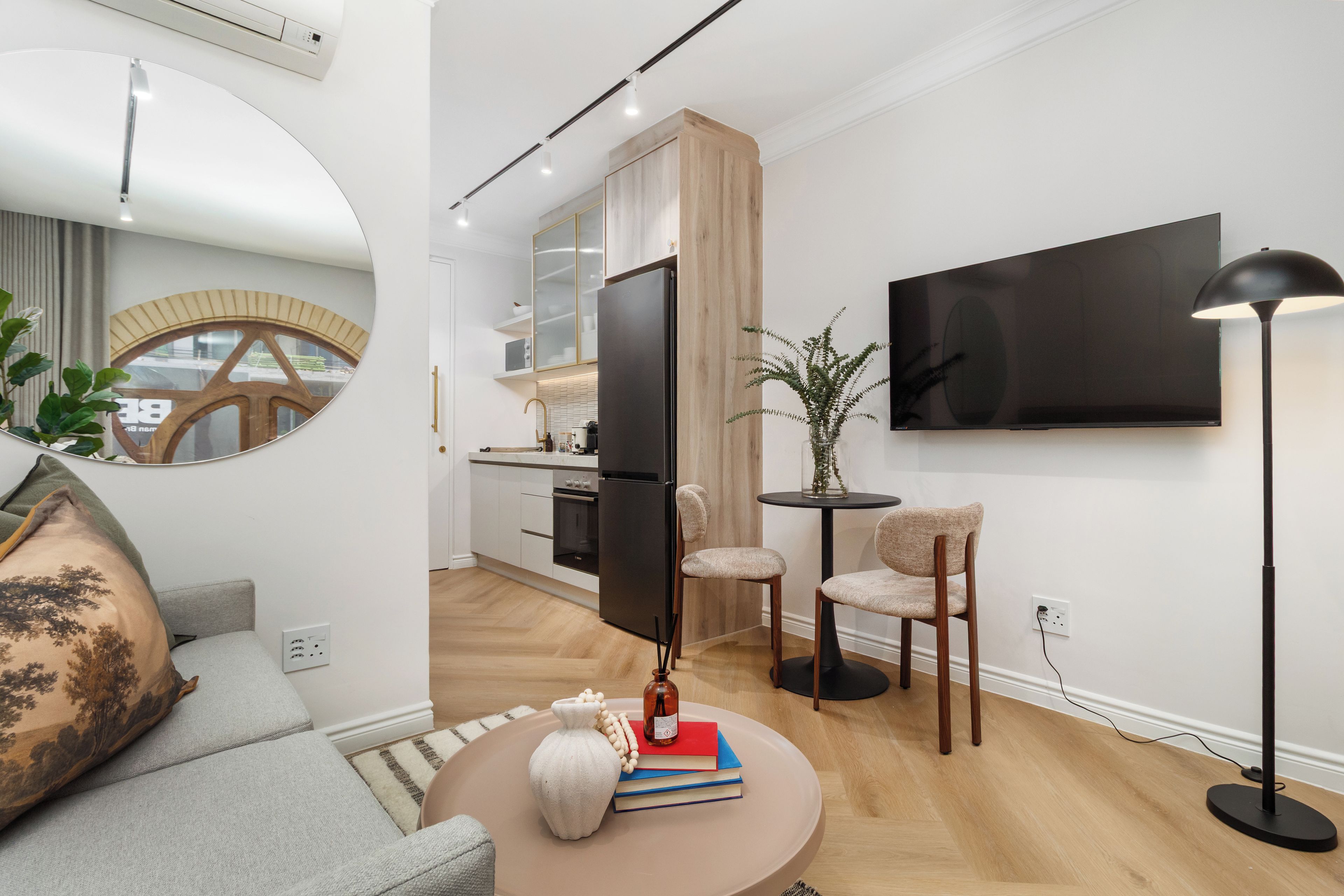 A modern living room with a sofa, a coffee table with books and a vase, a TV mounted on the wall, and a dining area with chairs and a table.