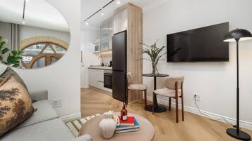 A modern living room with a sofa, a coffee table with books and a vase, a TV mounted on the wall, and a dining area with chairs and a table.