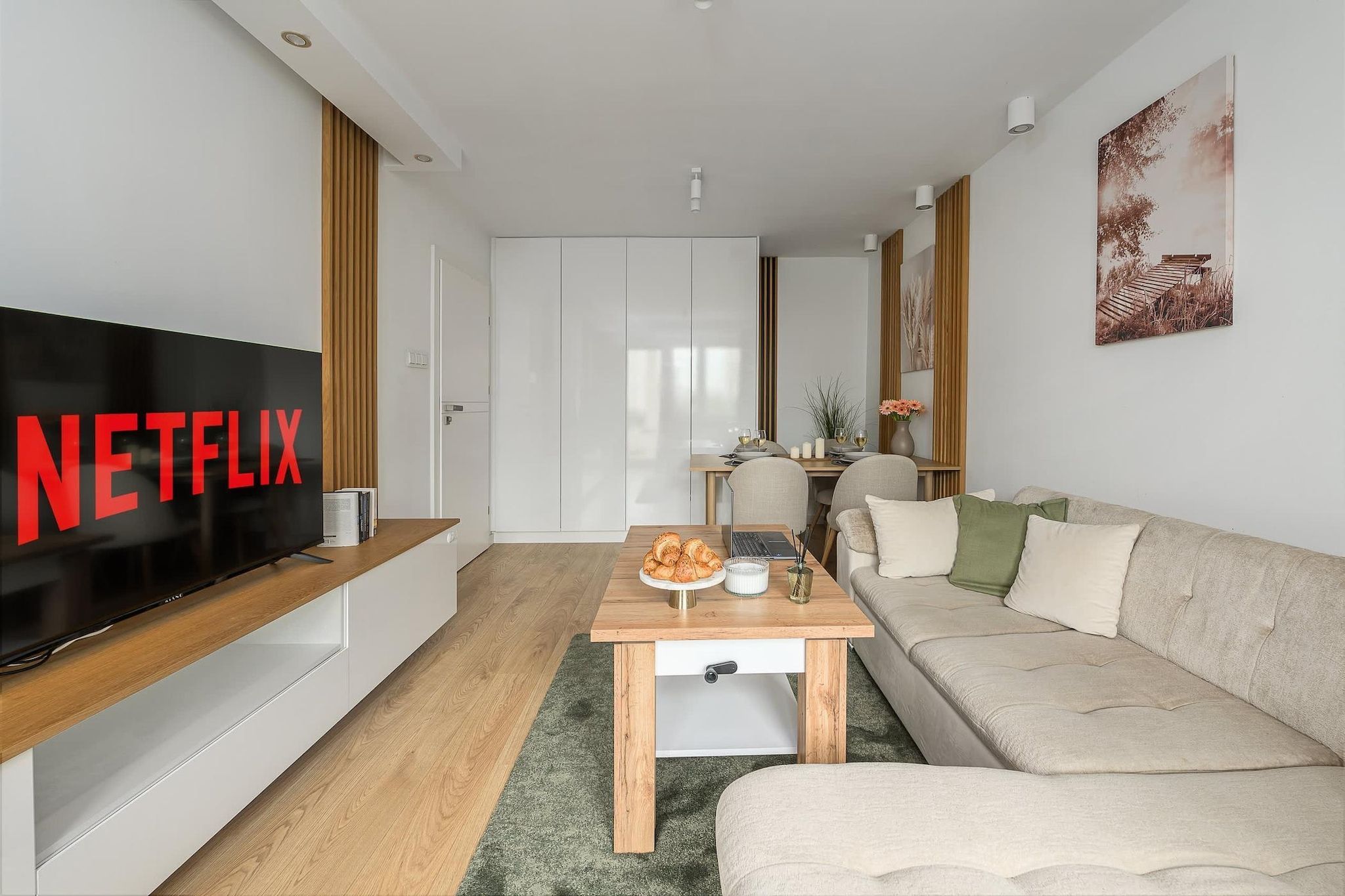 Een moderne woonkamer met een flatscreen televisie, een houten salontafel, een bank en een eethoek met een tafel en stoelen.