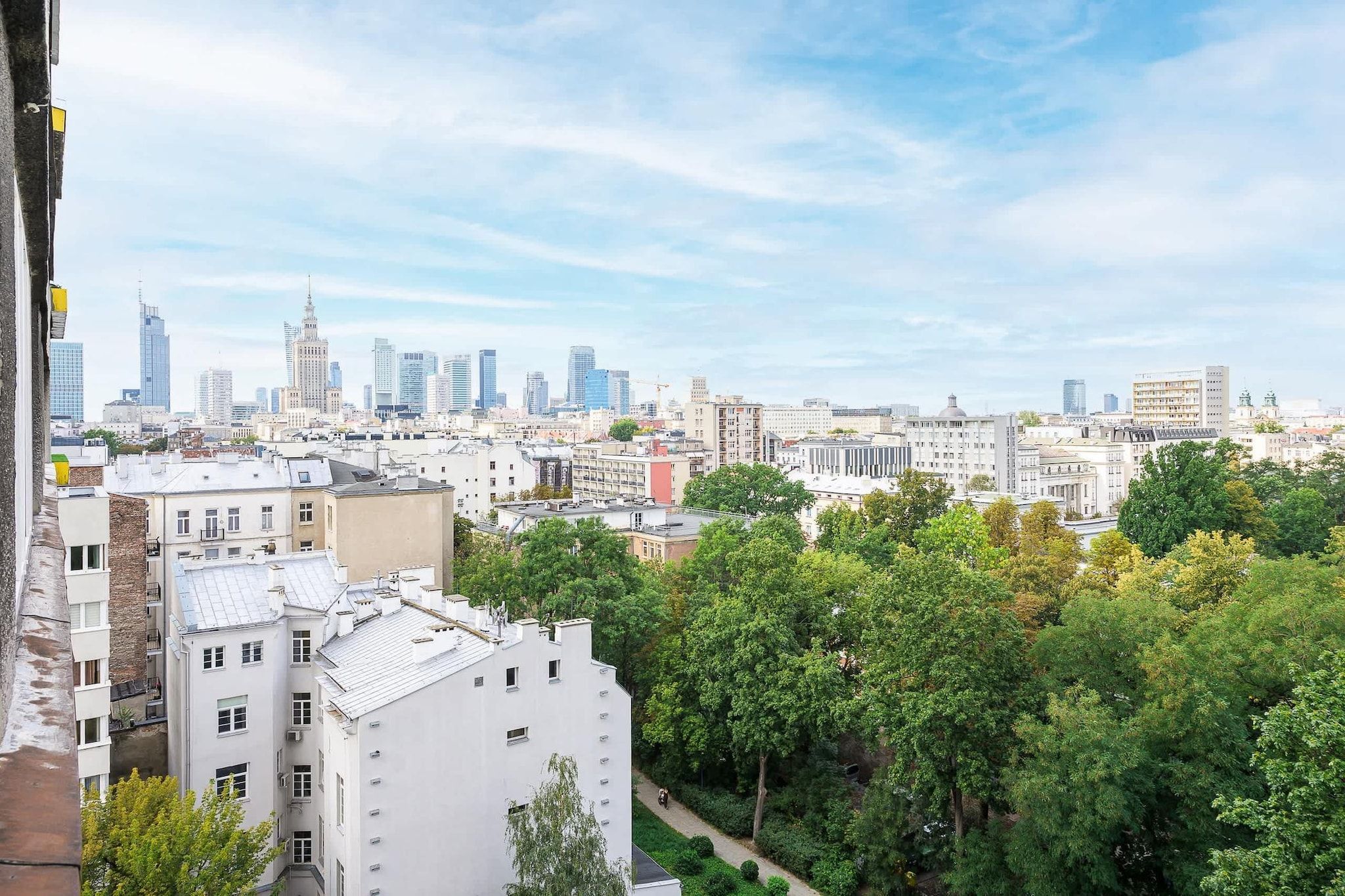 A cityscape with a mix of modern and historic buildings, dense urban greenery, and a clear sky.