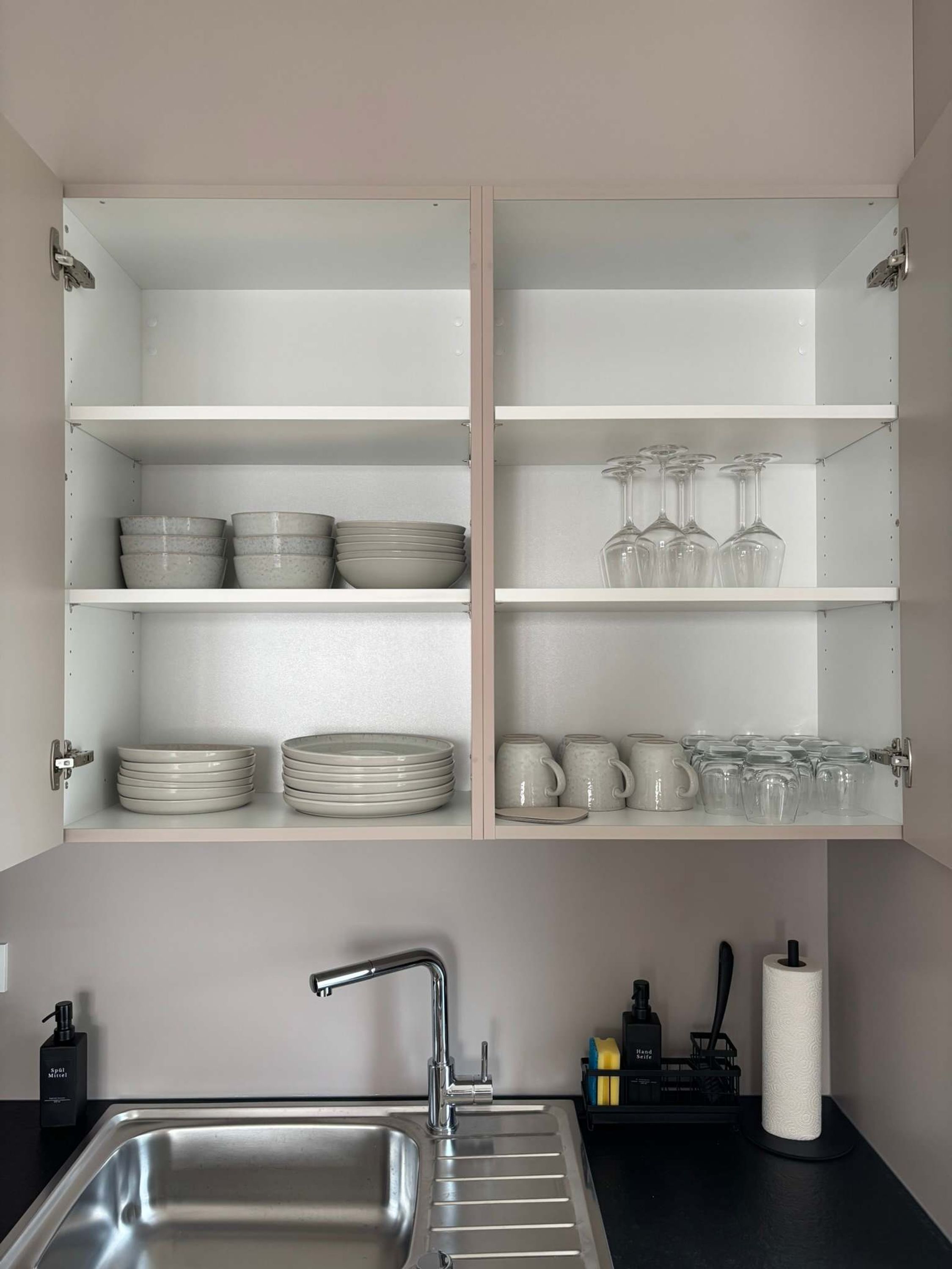 A kitchen with a stainless steel sink, open shelving with plates, bowls, glasses, and mugs.