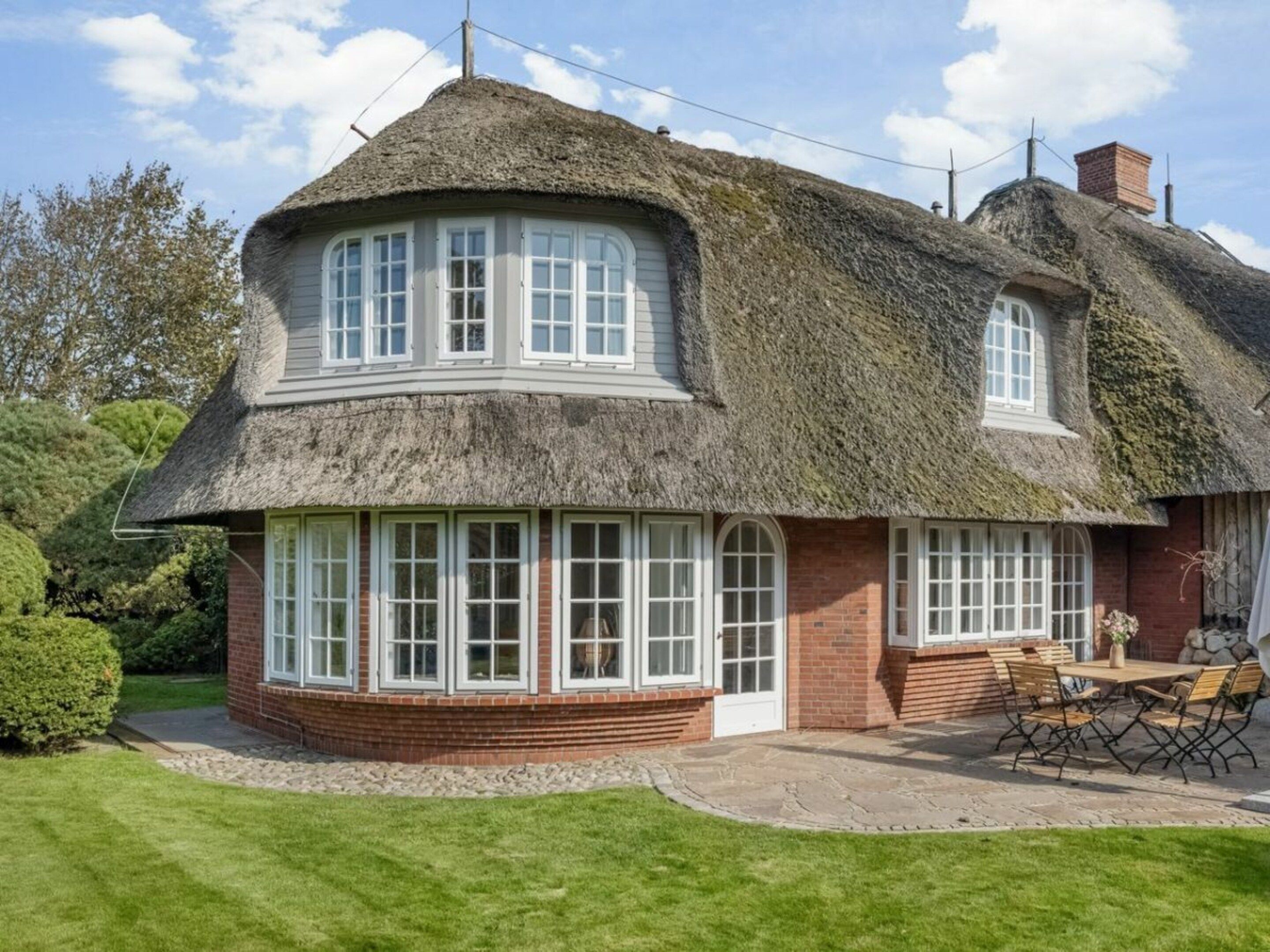 Une maison à toit de chaume, avec une base en briques, de grandes fenêtres et une terrasse aménagée avec du mobilier d’extérieur.