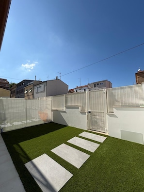 Un patio con césped artificial, una pared blanca y una reja.