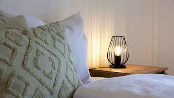 Un lit bien fait, agrémenté d’un plaid à motifs et de coussins, une table de chevet avec une lampe moderne, et une fenêtre ornée de rideaux légers.