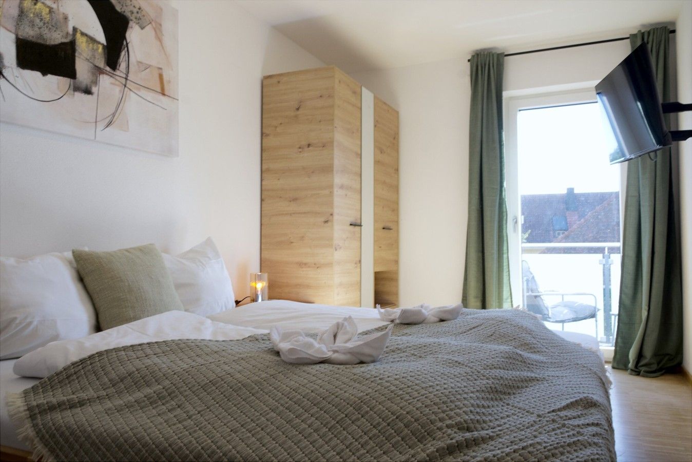 A hotel room with a bed, a wooden wardrobe, a window with curtains, and a flat-screen TV mounted on the wall.