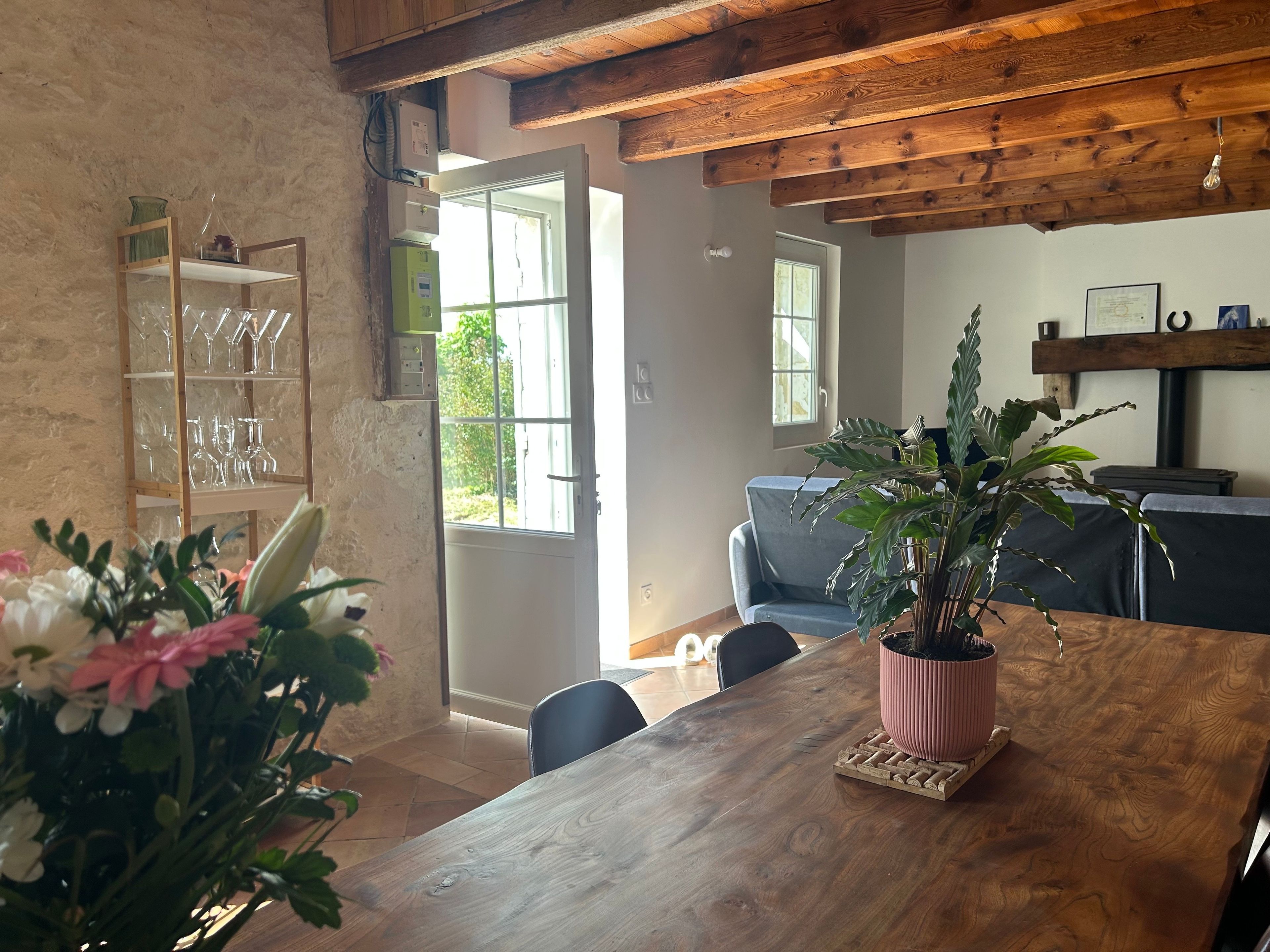 A wooden dining table with a potted plant, a glass display case with wine glasses, and a grey sofa in a room with exposed wooden beams and a window.