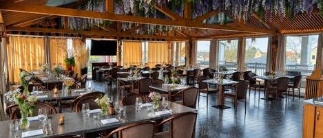 A spacious restaurant with multiple dining tables and chairs, a wooden ceiling, and hanging floral decorations.
