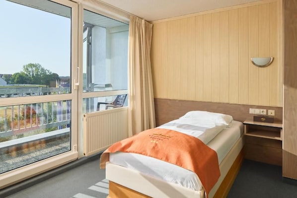 Habitación de hotel con cama, ventana con cortinas, cabecero de madera y mesita de noche pequeña.