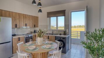 Een moderne keuken met houten kastjes, een eethoek met een ronde tafel en stoelen, en uitzicht op het landschap door een groot raam.