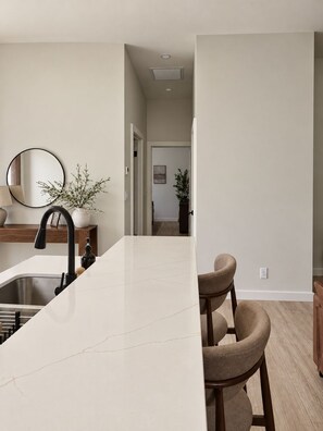 A modern kitchen with a white marble countertop, a black faucet, and two beige bar stools. - Farmhouse Suites - Plunge Pool, Sun Loungers, Grill, Walk to Beach, Beach Gear (Nassau)