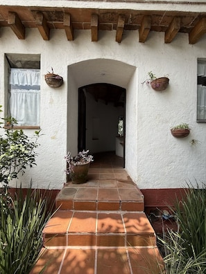 A building entrance with a tiled pathway leading to an archway, surrounded by potted plants and a window with curtains. - Casa del Viento · Vista al lago y terraza (Valle de Bravo)
