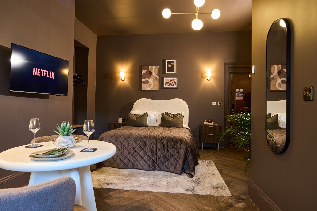 Habitación de hotel moderna con cama, mesita de noche, televisor montado en la pared y una mesa redonda con piña y copas de vino.