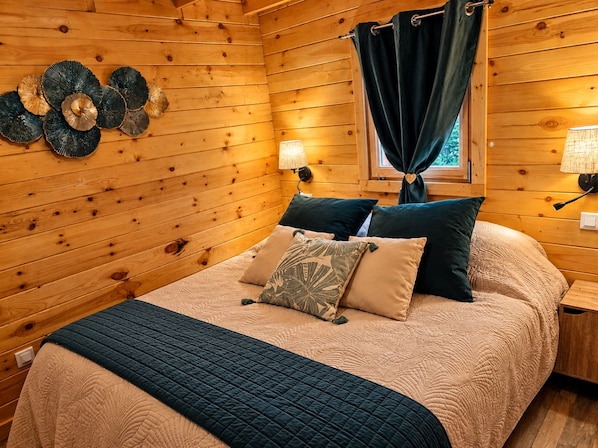Un dormitorio de cabaña de madera con una cama, almohadas, un edredón acolchado, una lámpara de pared y una ventana con cortinas.