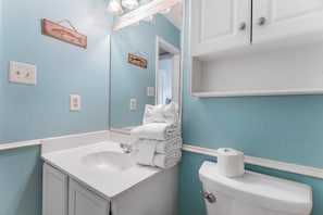 Un baño con lavamanos, inodoro y espejo. Hay toallas apiladas sobre el lavamanos y un rollo de papel higiénico en el inodoro.