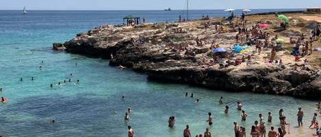 A crowded beach with people swimming and sunbathing, rocky terrain, and a clear view of the sea.