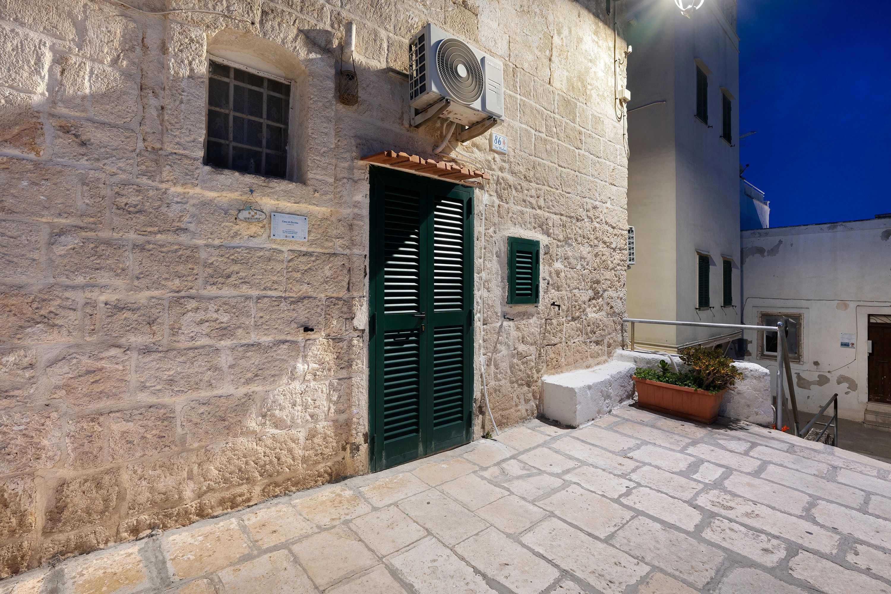 A stone building with a green door, a sign indicating room number 86, and a small planter with plants.