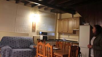 Un salon chaleureux avec un canapé, une table à manger en bois et des chaises. Une personne se tient près de l’espace cuisine, où se trouvent des placards et un four à micro-ondes.