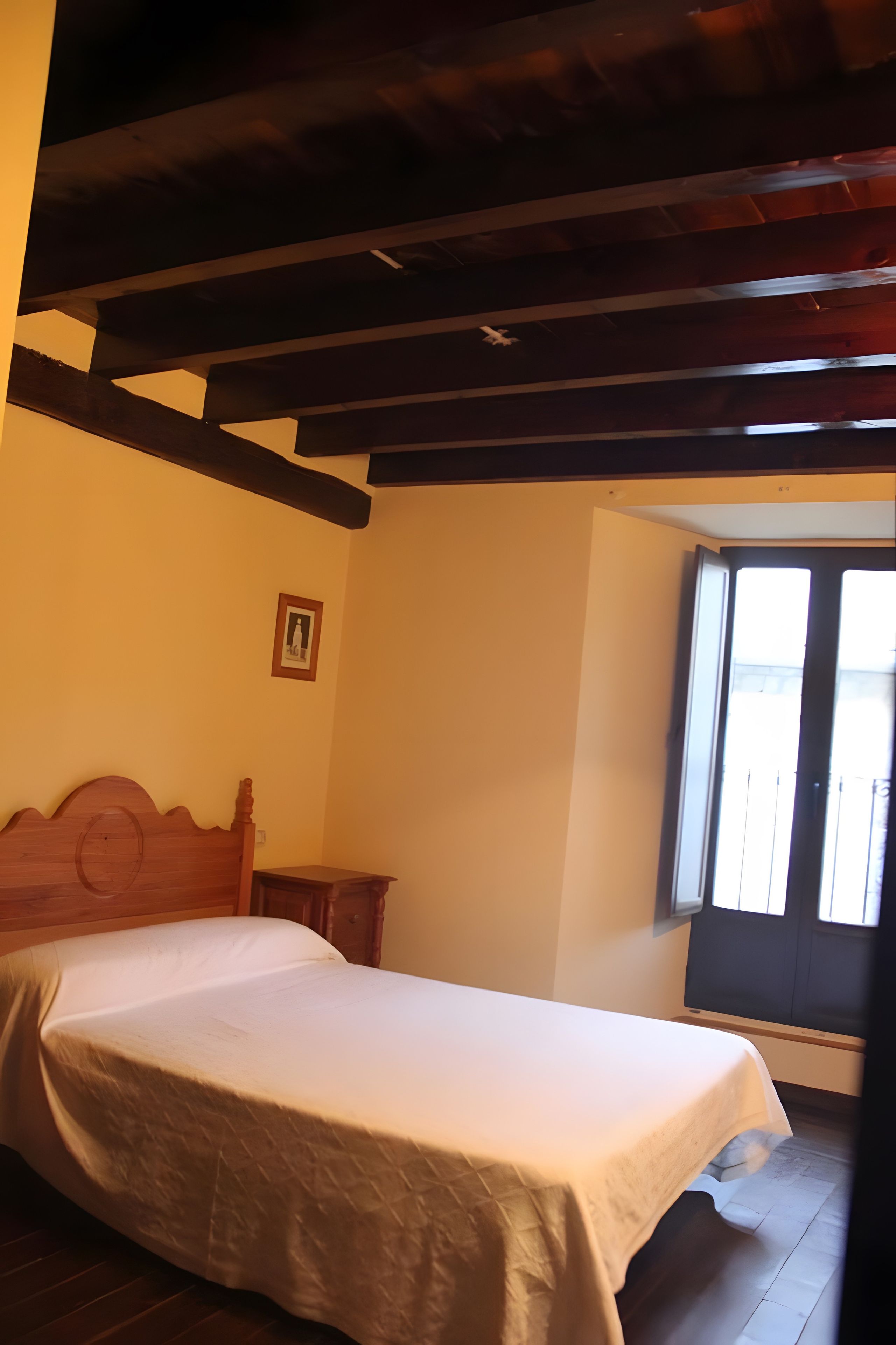 Une chambre à coucher avec un lit, une tête de lit en bois, une petite table de chevet et une fenêtre avec des stores.