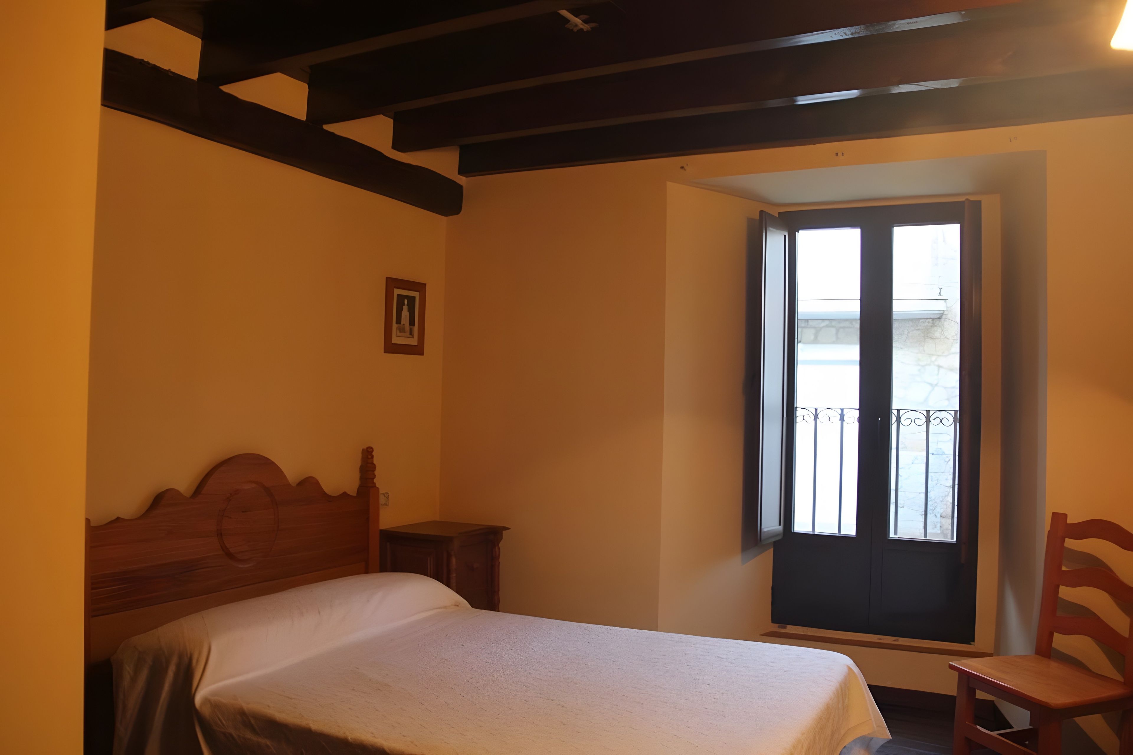 A bedroom with a bed, a chair, a nightstand, a window with a view, and a picture on the wall.