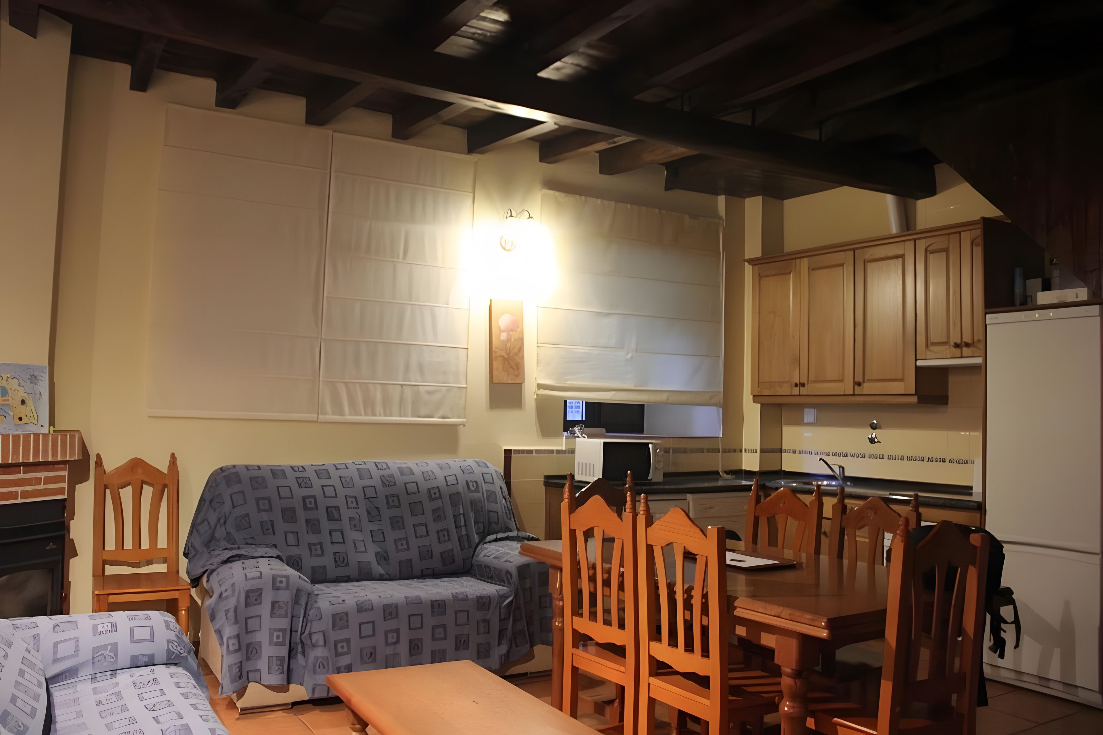 A living room with a sofa, a wooden dining table with chairs, a kitchen area with cabinets and appliances, and a ceiling with exposed beams.