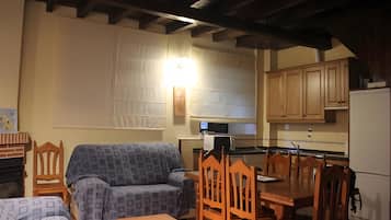 A living room with a sofa, a wooden dining table with chairs, a kitchen area with cabinets and appliances, and a ceiling with exposed beams.