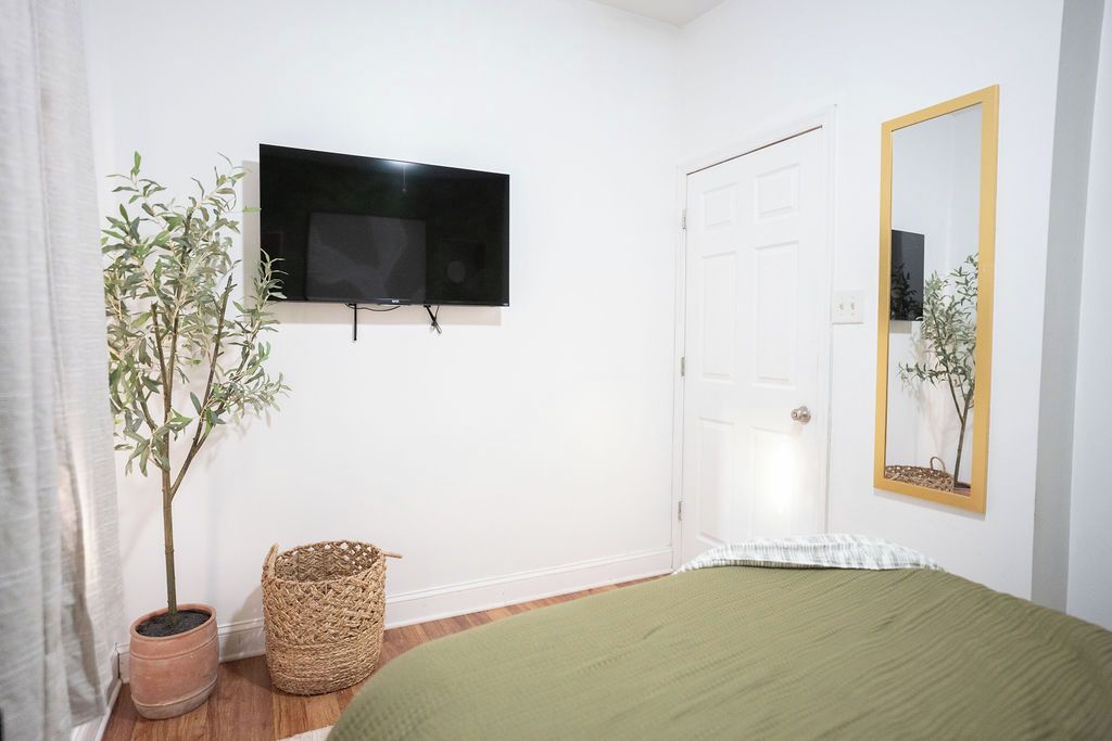 Un dormitorio con cama, televisor montado en la pared, puerta, espejo y planta en maceta.