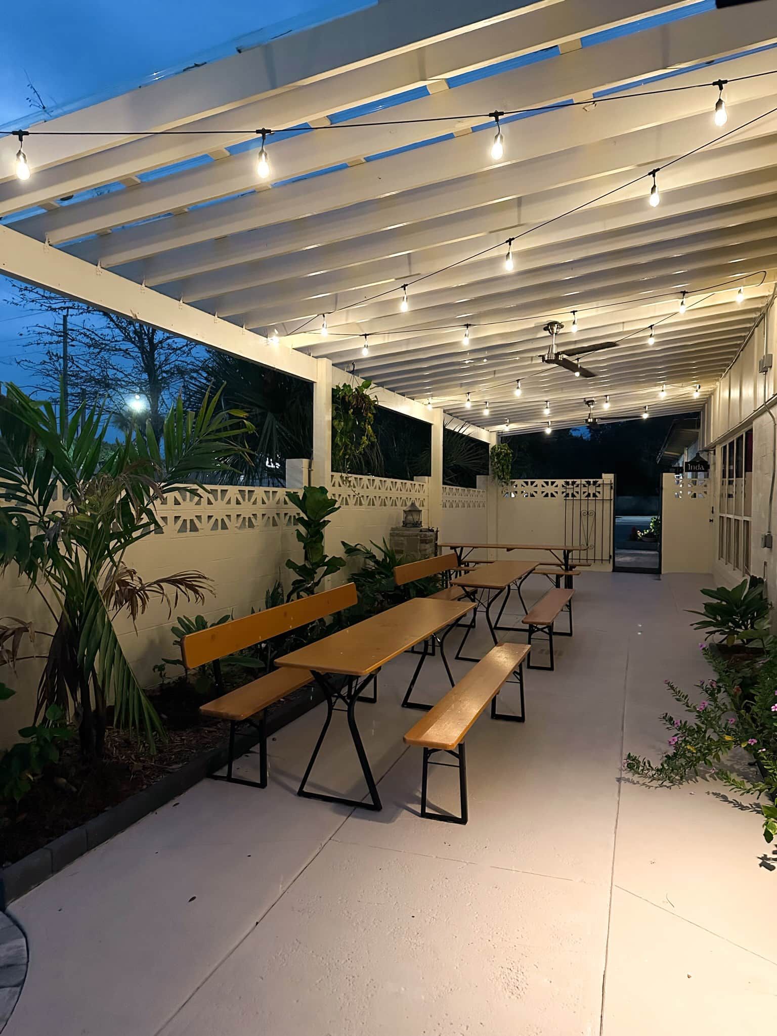 An outdoor patio area with multiple wooden benches and tables, a ceiling fan, and string lights.
