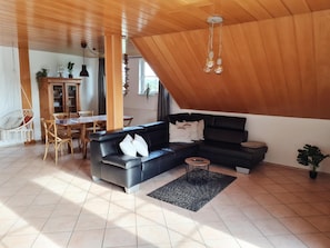 A modern living room with a black sectional sofa, a dining table with chairs, and a hanging light fixture. - Apartment 'Am Lutherweg 1521' with Balcony and Wi-Fi (Breitenbach am Herzberg)